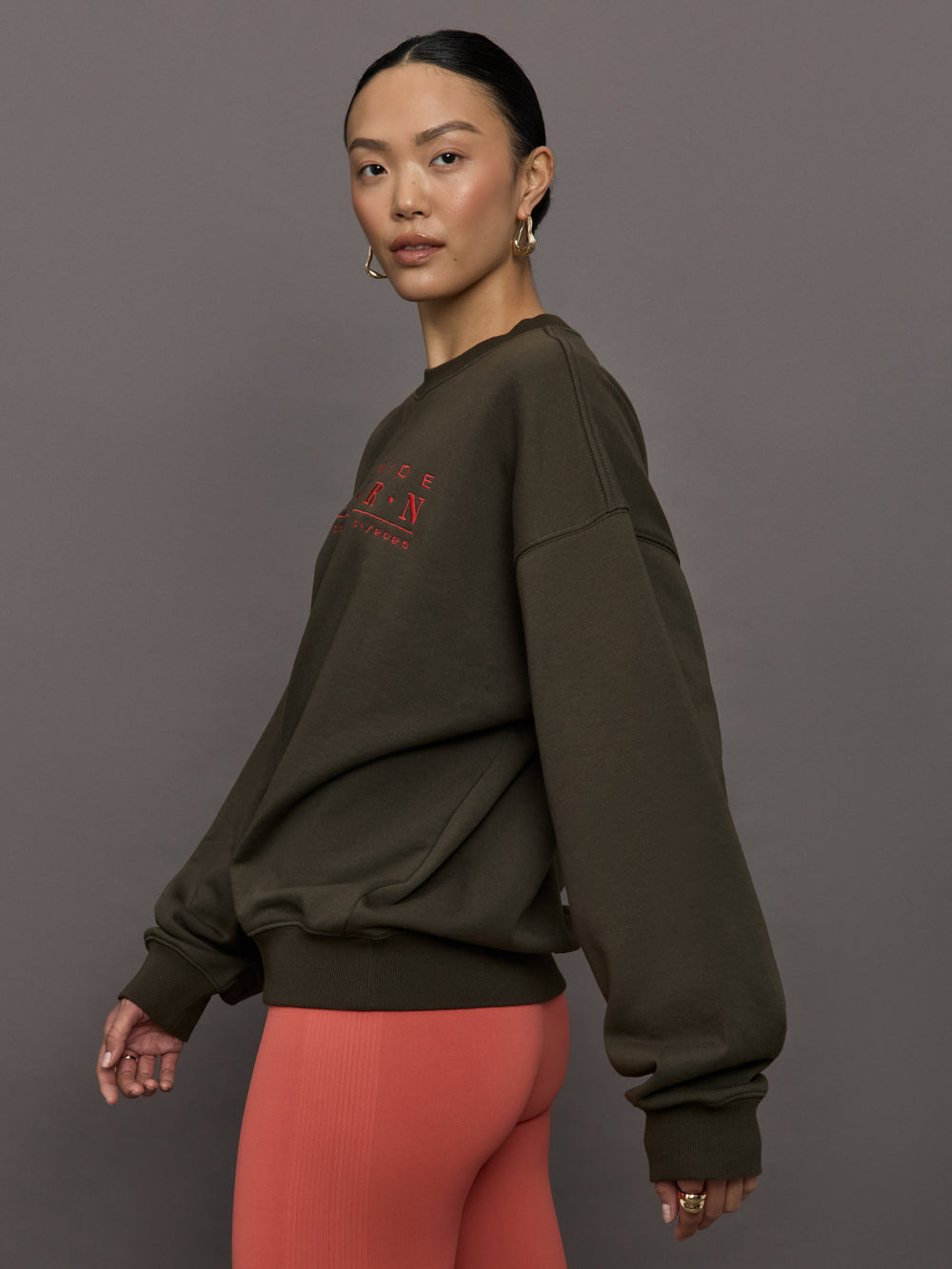 Model wears an oversized brown crewneck sweatshirt with the words 'Worldwide Modern' printed in coral coloring across the chest.