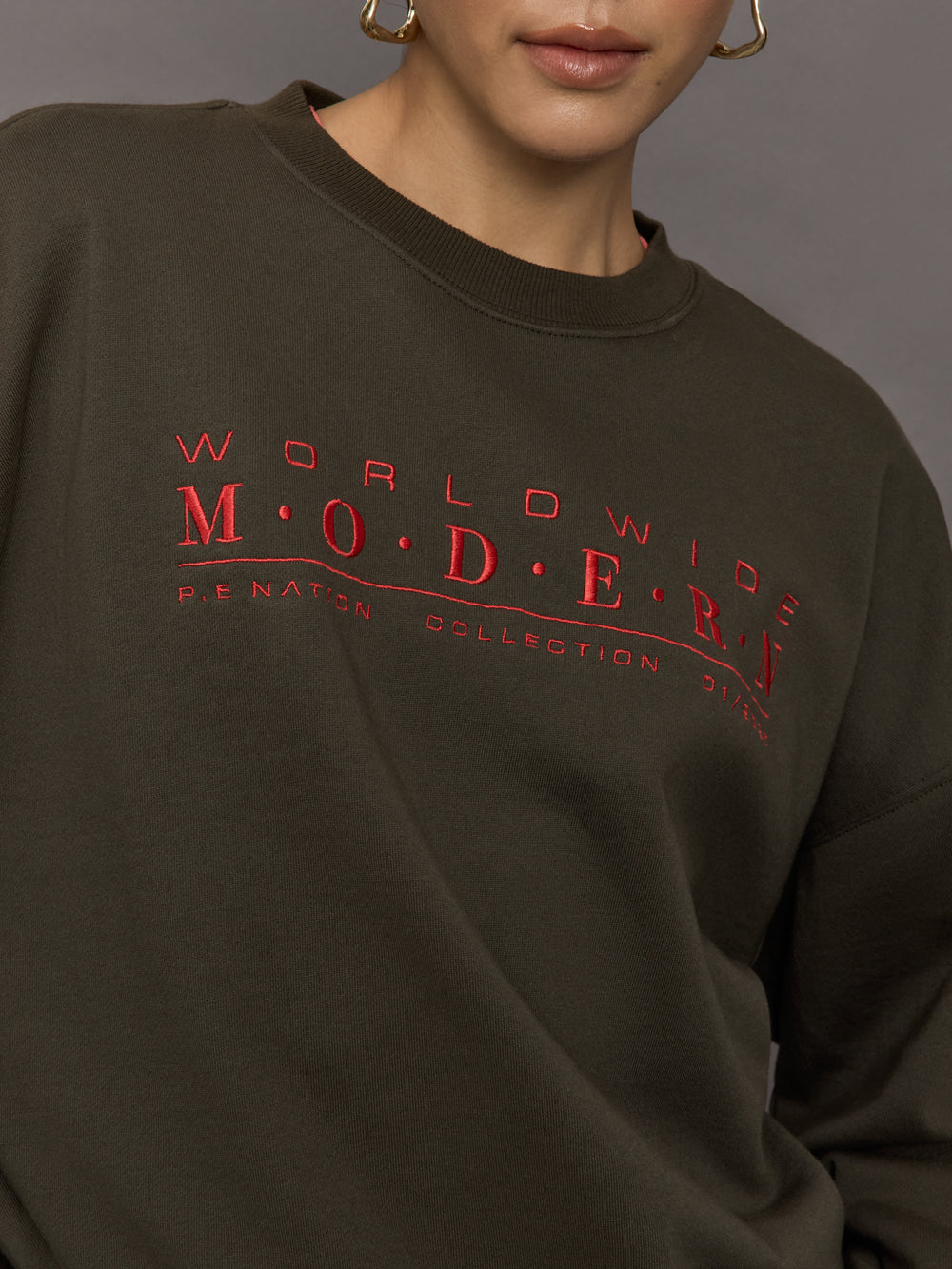 Model wears an oversized brown crewneck sweatshirt with the words 'Worldwide Modern' printed in coral coloring across the chest.
