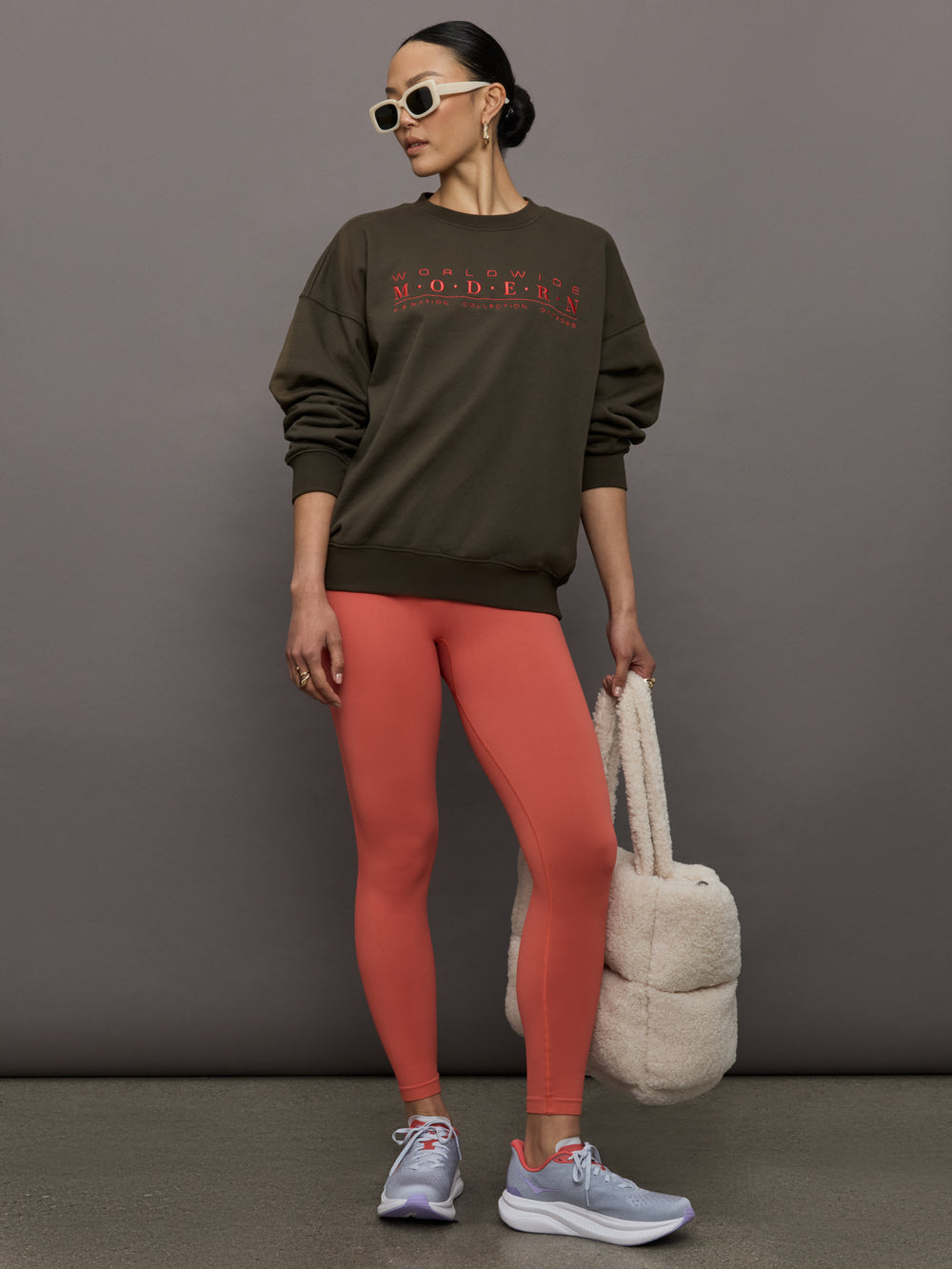 Model wears an oversized brown crewneck sweatshirt with the words 'Worldwide Modern' printed in coral coloring across the chest.