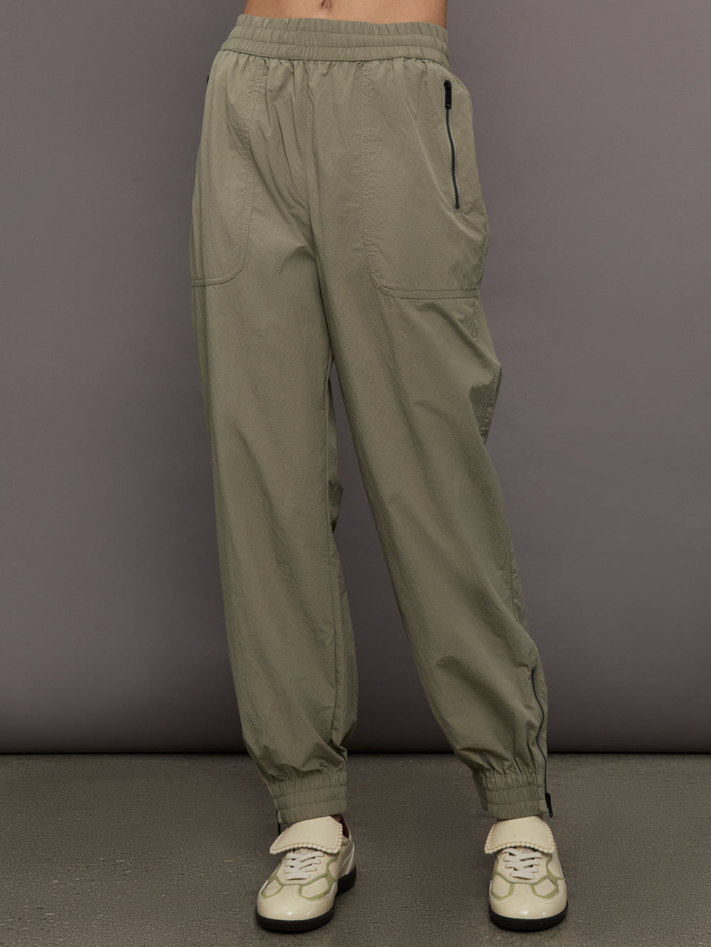 Model wears khaki track pant with large front pockets and cuffed ankles. 