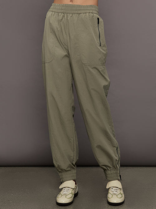 Model wears khaki track pant with large front pockets and cuffed ankles. 