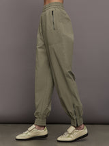 Model wears khaki track pant with large front pockets and cuffed ankles. 