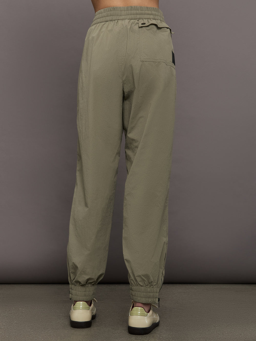 Model wears khaki track pant with large front pockets and cuffed ankles. 