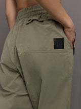 Model wears khaki track pant with large front pockets and cuffed ankles. 