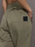 Model wears khaki track pant with large front pockets and cuffed ankles. 