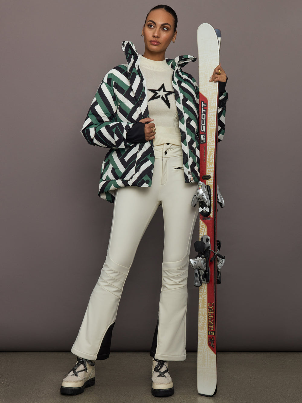 Full body view of model wearing Aurora High Waist Ski Pant Snow White styled with star sweater and printed ski jacket.
