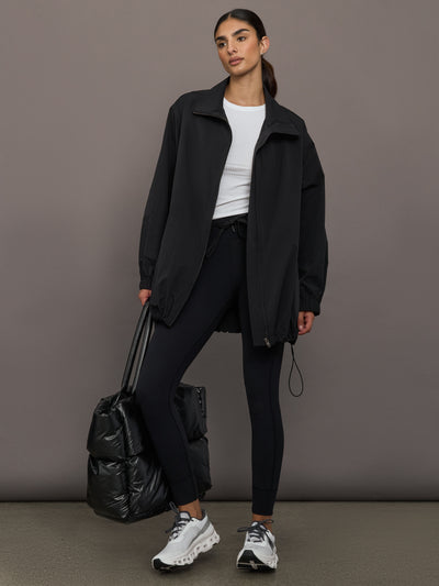 Woman wearing a black coat and black pants, holding a black bag against a gray background