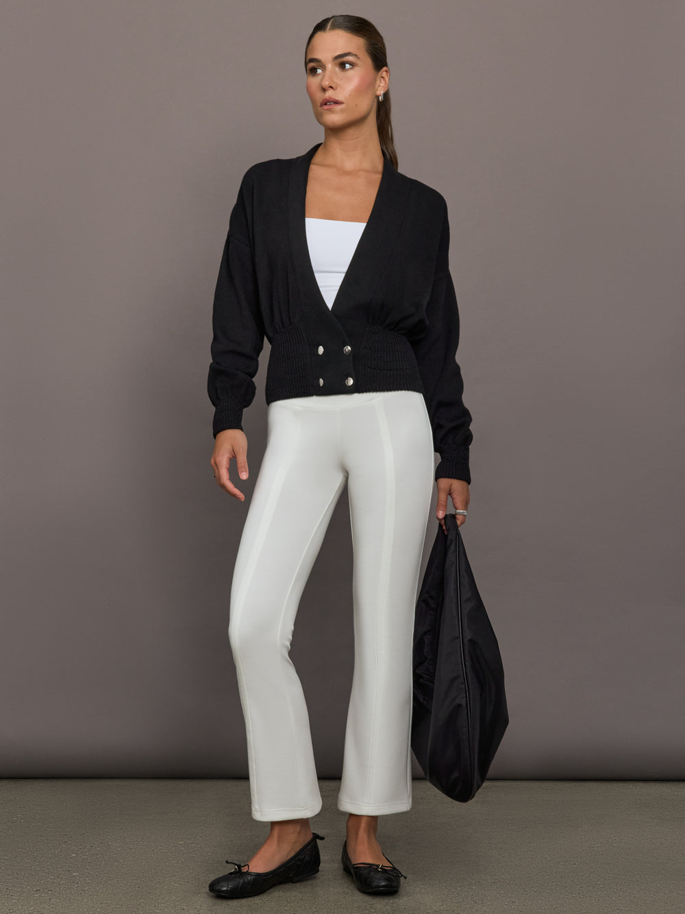 Model is wearing a black ribbed-waist cardigan with deep V-neckline and button detail, styled with white straight-leg pants, black flats, and a black shoulder bag, full body view.