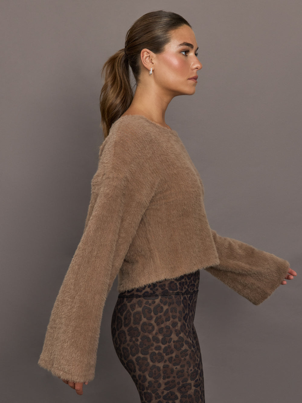 Model is wearing a taupe plush long-sleeve top with wide bell sleeves and relaxed fit, styled with leopard print leggings, side view.
