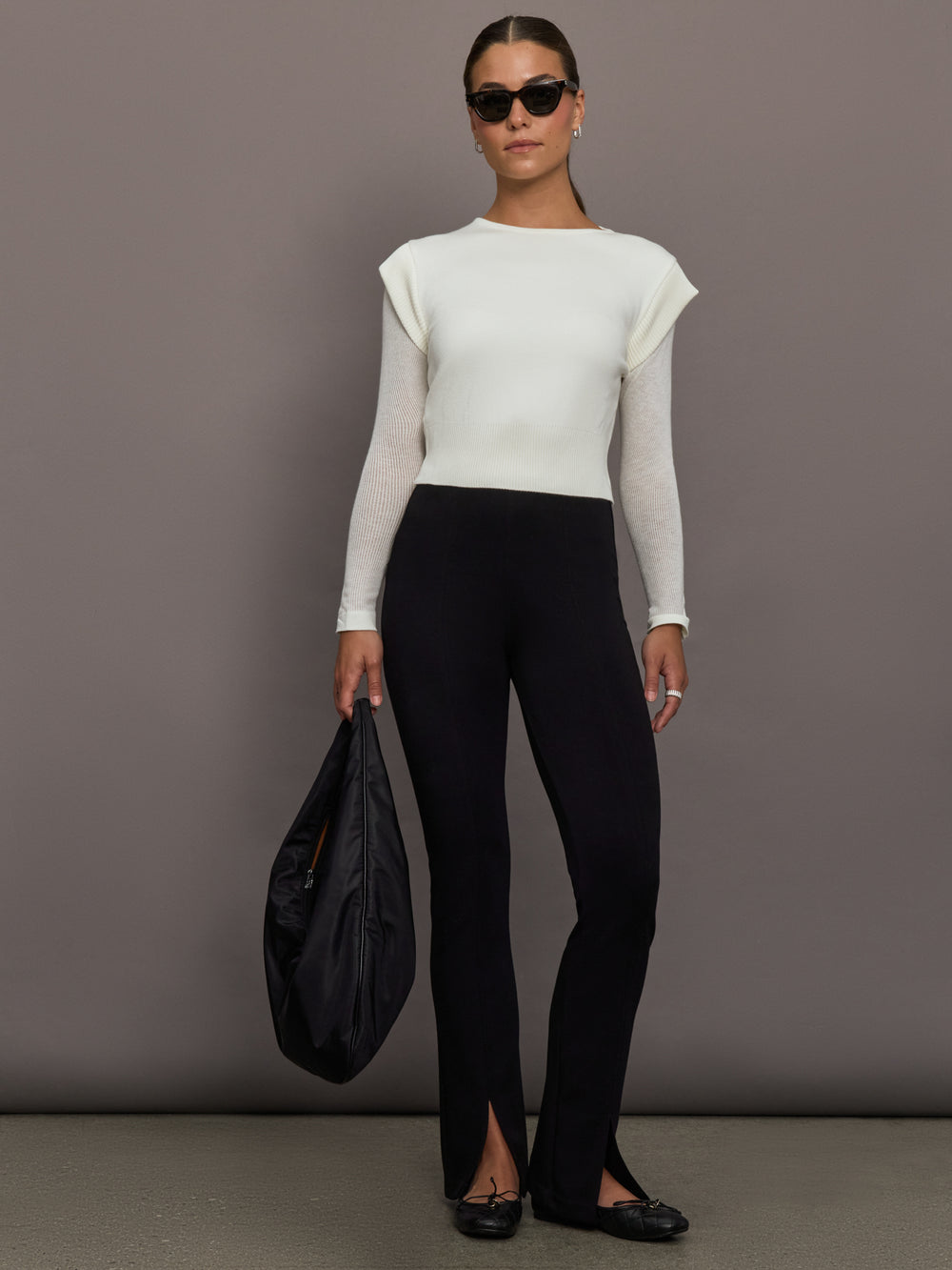 Model is wearing a white crew-neck sweater with sheer mesh long sleeves, styled with black split-hem pants, black flats, oversized sunglasses, and a black shoulder bag, full body view.