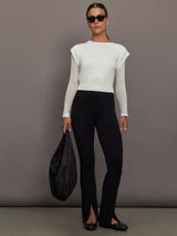 Model is wearing a white crew-neck sweater with sheer mesh long sleeves, styled with black split-hem pants, black flats, oversized sunglasses, and a black shoulder bag, full body view.