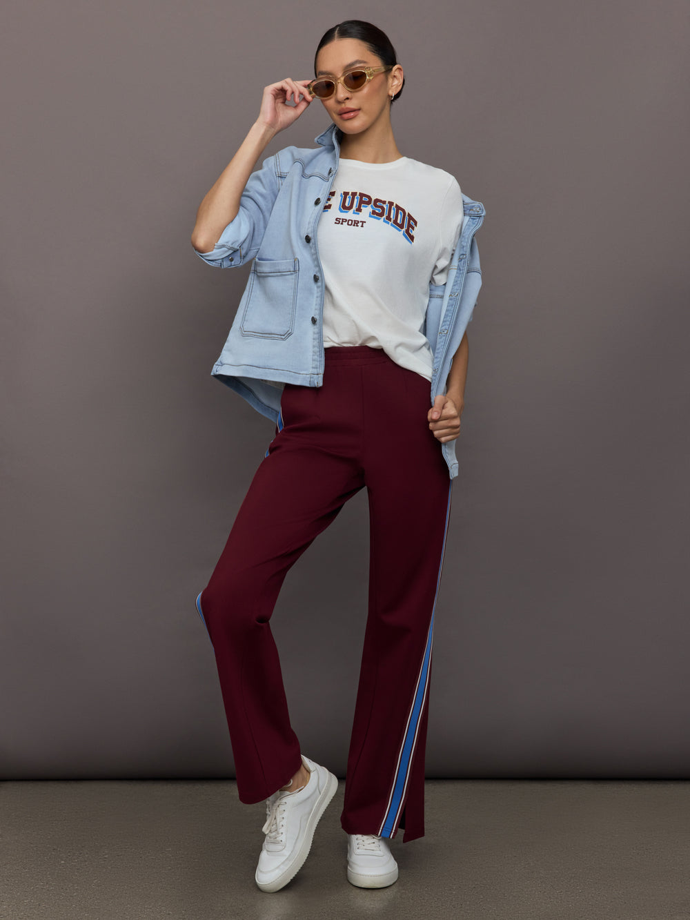 Woman wearing a denim jacket, white graphic t-shirt, maroon pants with a blue stripe, and white sneakers against a gray background.