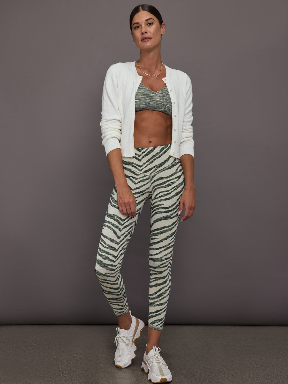 Woman wearing a white cardigan, sports bra, and zebra-striped leggings against a gray background