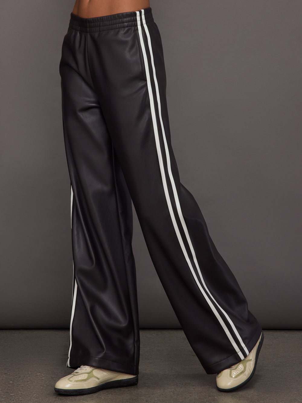 Model wears a pair of dark brown vegan leather track pants with white stipes down side. 
