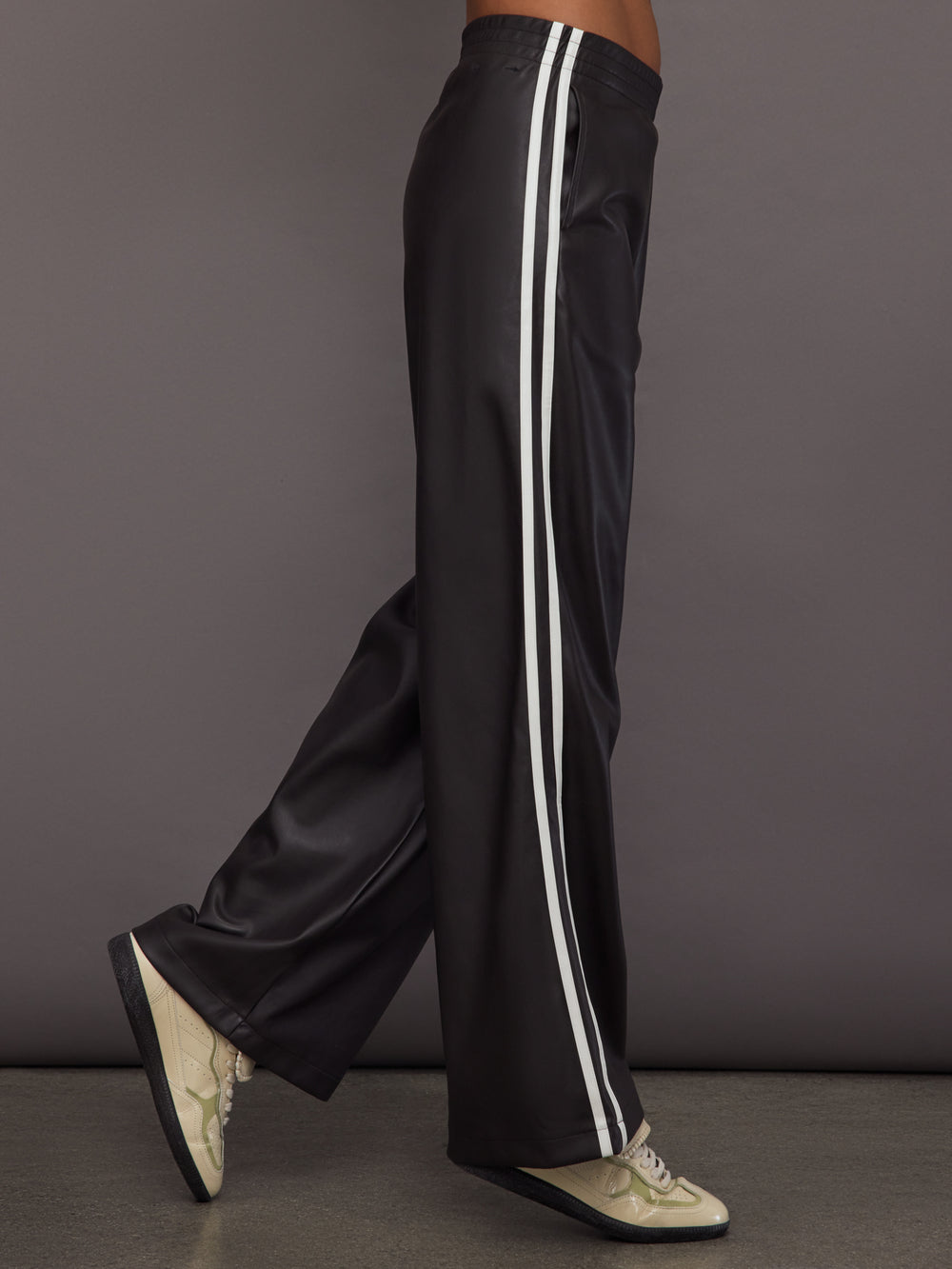 Model wears a pair of dark brown vegan leather track pants with white stipes down side. 