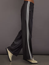 Model wears a pair of dark brown vegan leather track pants with white stipes down side. 