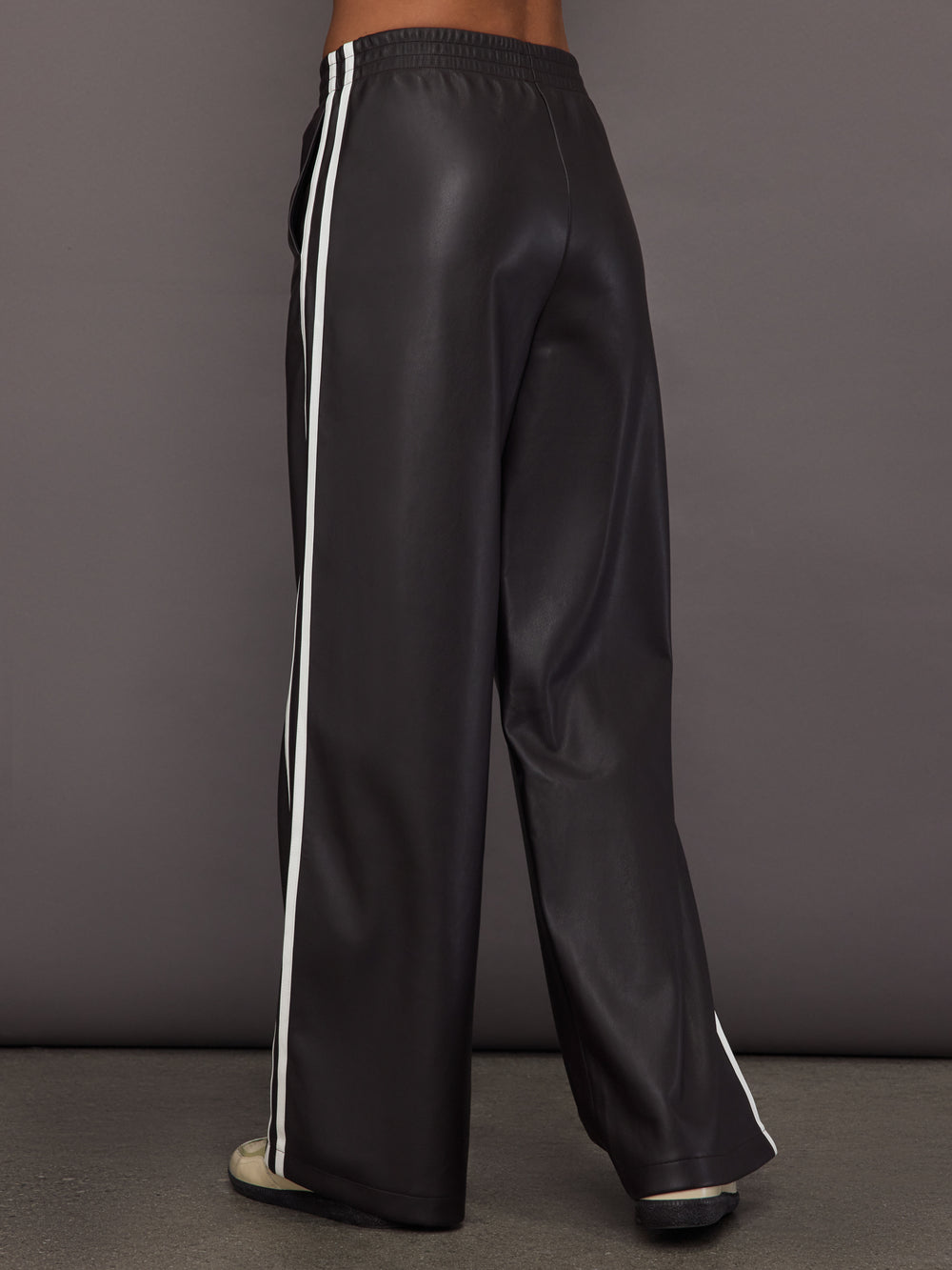 Model wears a pair of dark brown vegan leather track pants with white stipes down side. 