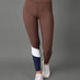 A model wearing a brown high-waisted activewear set consisting of a sports bra and leggings, featuring a color-blocked design on the lower leg with white and navy blue panels, paired with white sneakers against a solid gray studio background.