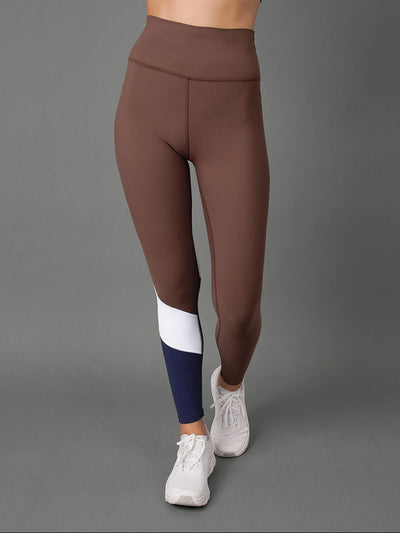 A model wearing a brown high-waisted activewear set consisting of a sports bra and leggings, featuring a color-blocked design on the lower leg with white and navy blue panels, paired with white sneakers against a solid gray studio background.