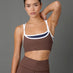 A woman modeling a matching brown activewear set consisting of a sports bra with white and navy trim and high-waisted leggings, set against a solid gray studio background.