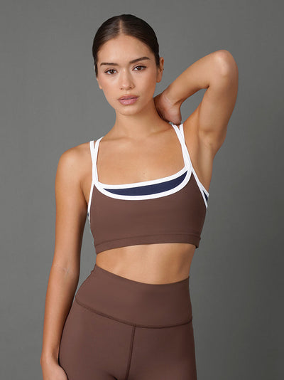 A woman modeling a matching brown activewear set consisting of a sports bra with white and navy trim and high-waisted leggings, set against a solid gray studio background.