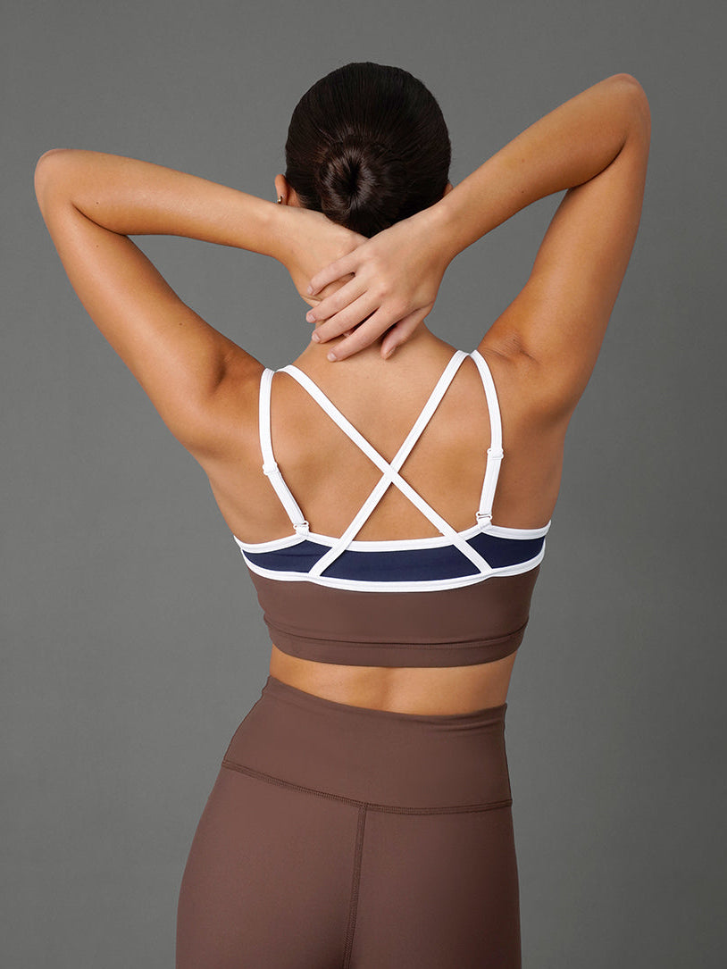 A woman shown from the back wearing a brown and navy sports bra with white crisscross straps and matching high-waisted brown leggings, posed against a solid gray studio background.