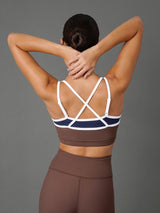 A woman shown from the back wearing a brown and navy sports bra with white crisscross straps and matching high-waisted brown leggings, posed against a solid gray studio background.