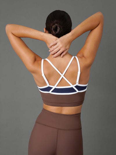 A woman shown from the back wearing a brown and navy sports bra with white crisscross straps and matching high-waisted brown leggings, posed against a solid gray studio background.