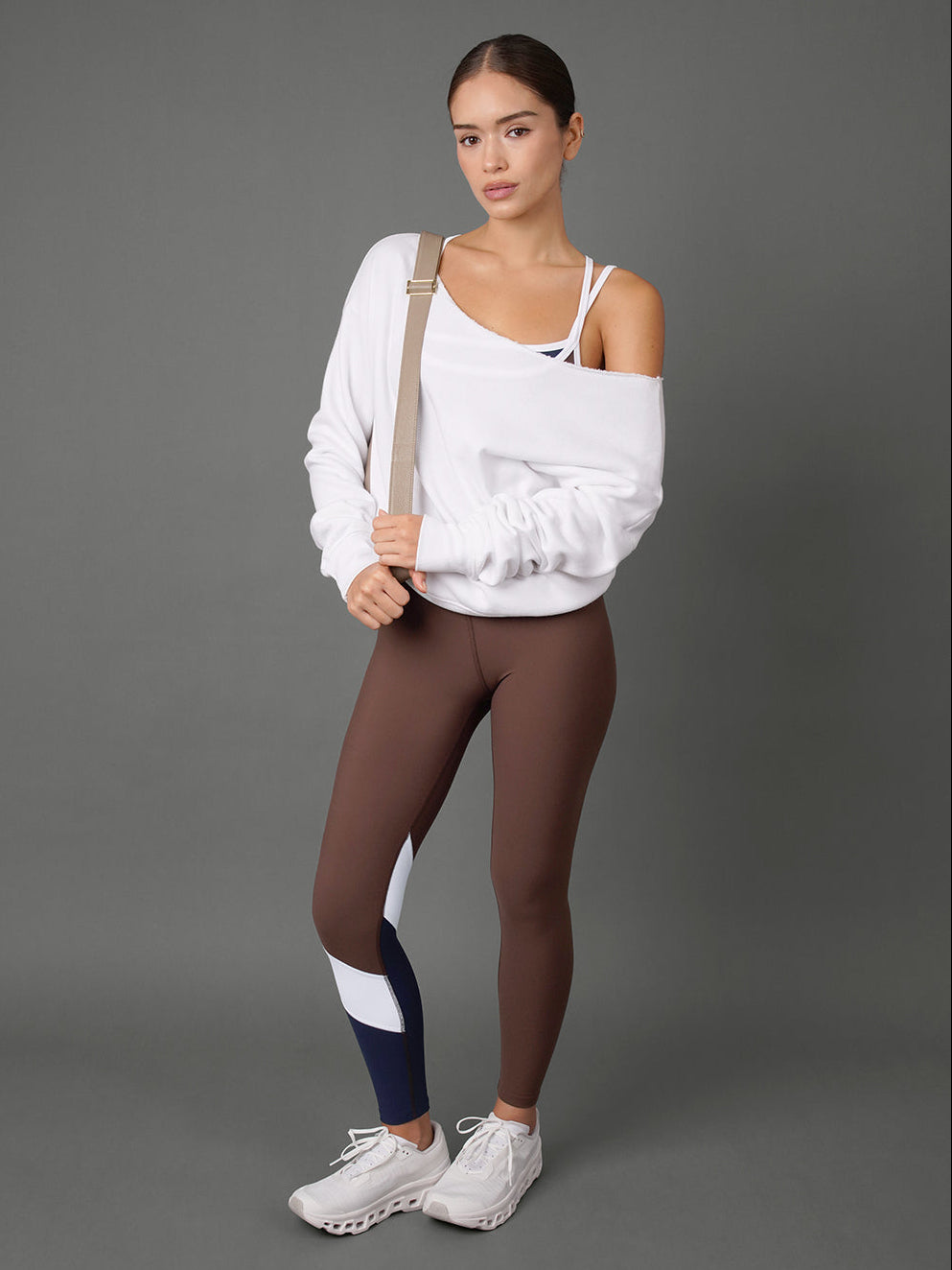 A model stands against a solid gray studio background wearing a white off-the-shoulder sweatshirt, brown leggings with white and navy color-blocked panels on the lower leg, and white sneakers.