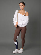 A model stands against a solid gray studio background wearing a white off-the-shoulder sweatshirt, brown leggings with white and navy color-blocked panels on the lower leg, and white sneakers.