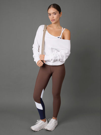 A model stands against a solid gray studio background wearing a white off-the-shoulder sweatshirt, brown leggings with white and navy color-blocked panels on the lower leg, and white sneakers.