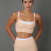 A woman modeling a matching beige athletic set consisting of a scoop-neck sports bra and high-waisted leggings, featuring a white underband on the sports bra and a white waistband on the leggings, set against a solid gray studio background.