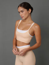 A woman modeling a matching beige and white two-piece athletic set, consisting of a scoop-neck sports bra and high-waisted leggings, against a solid dark gray studio background.