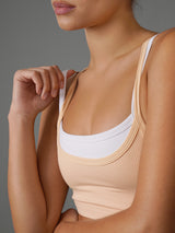 Close-up of a woman wearing a ribbed scoop-neck tank top in a light peach-beige color, layered over a white underband, against a solid gray studio background.