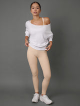 A woman modeling a white off-the-shoulder long-sleeve top and beige leggings with white sneakers, standing against a solid gray studio background.