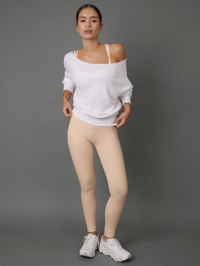 A woman modeling a white off-the-shoulder long-sleeve top and beige leggings with white sneakers, standing against a solid gray studio background.