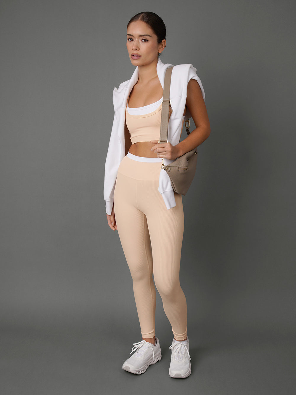 A woman modeling a matching beige activewear set consisting of high-waisted leggings and a sports bra, styled with a white zip-up hoodie, a beige shoulder bag, and white sneakers against a solid gray studio background.
