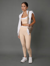 A woman modeling a matching beige activewear set consisting of high-waisted leggings and a sports bra, styled with a white zip-up hoodie, a beige shoulder bag, and white sneakers against a solid gray studio background.