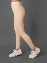A model shown from the side wearing a matching beige activewear set consisting of a sports bra and high-waisted leggings, paired with white athletic sneakers, against a solid dark gray studio background.