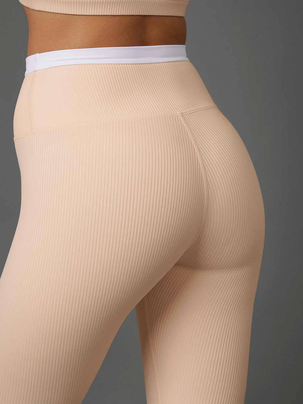 Close-up of a model wearing ribbed high-waisted leggings in a light peach-beige color, showing the back and side fit against a solid dark gray background.