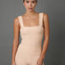 A woman modeling a beige tank with a square neckline and wide shoulder straps, paired with matching high-waisted leggings, against a solid dark gray studio background.