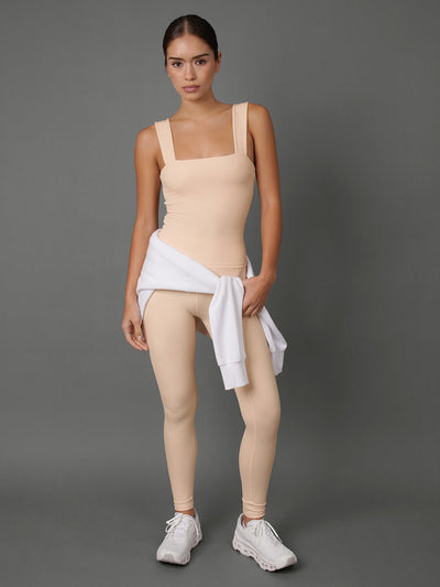 A model stands against a solid gray studio background wearing a beige tank, matching leggings, and white sneakers, with a white sweatshirt tied around her waist.