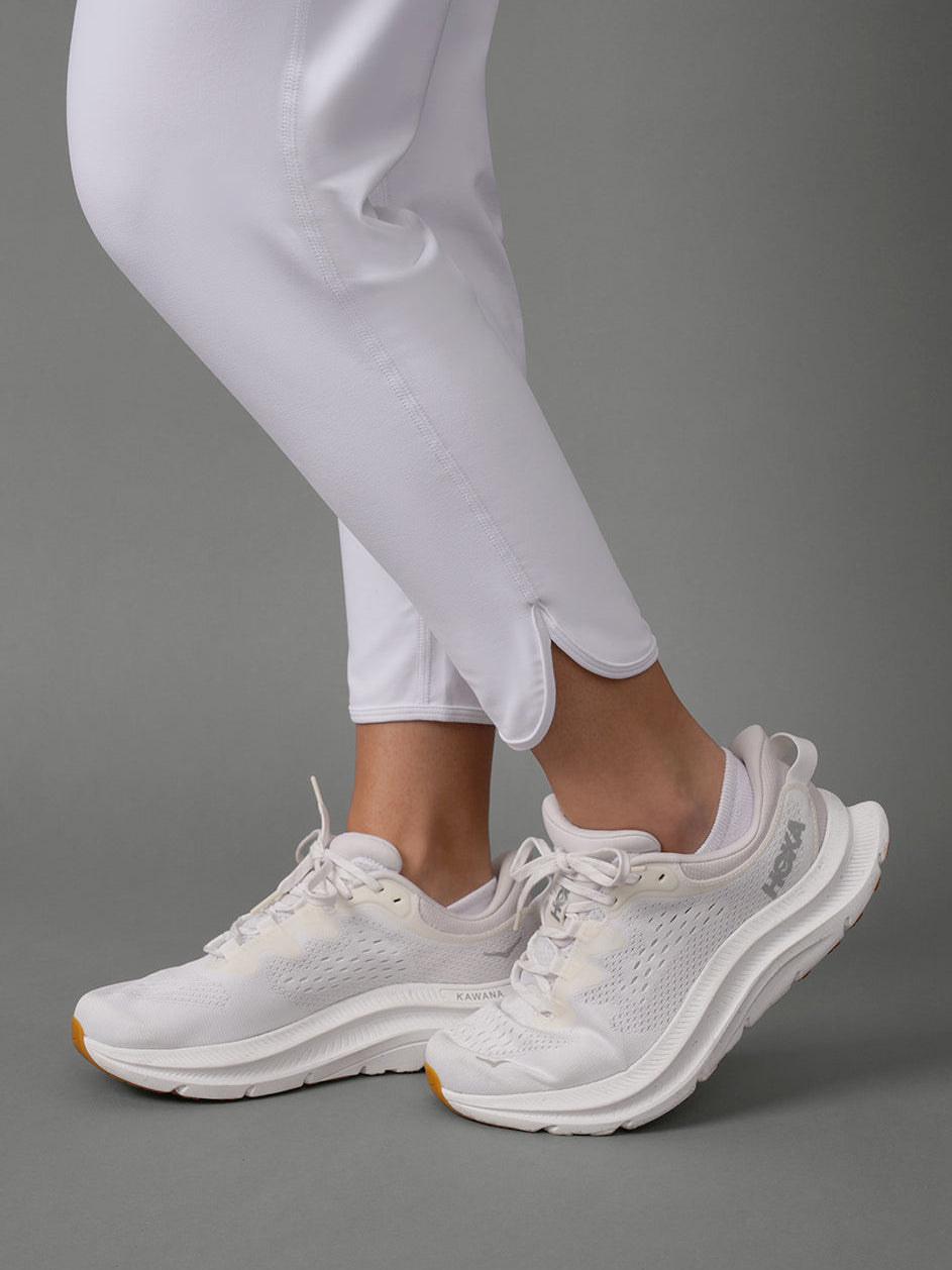 White athletic sneakers with a chunky layered sole, shown on a model wearing white pants against a solid gray studio background.