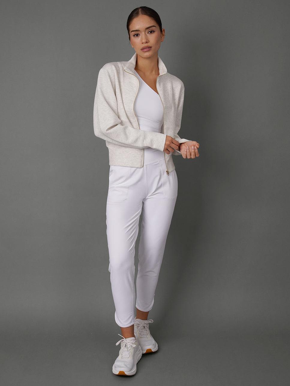 A woman modeling a white athletic tracksuit consisting of a zip-up jacket, a matching top, and slim-fit pants, paired with white sneakers, against a solid gray studio background.