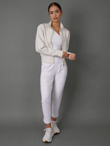 A woman modeling a white athletic tracksuit consisting of a zip-up jacket, a matching top, and slim-fit pants, paired with white sneakers, against a solid gray studio background.