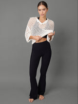 A woman stands against a plain gray studio background wearing a white open-knit cropped wrap top and black pants.
