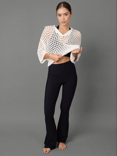 A woman stands against a plain gray studio background wearing a white open-knit cropped wrap top and black pants.