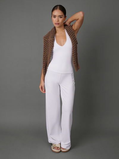 A woman modeling a white sleeveless top and matching white wide-leg pants, styled with a brown open-knit vest draped over her shoulders, against a solid gray studio background.