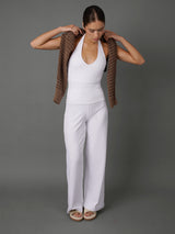 A woman modeling a white sleeveless top and matching white wide-leg pants, holding a brown open-knit cardigan over her shoulders against a solid gray studio background.