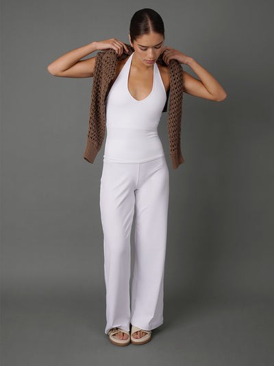 A woman modeling a white sleeveless top and matching white wide-leg pants, holding a brown open-knit cardigan over her shoulders against a solid gray studio background.